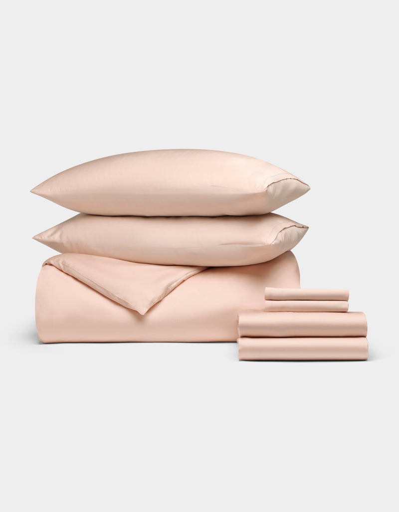The Bamboo Bedding Ultimate Bundle by Cozy Earth, in light peach, features two stacked pillows, a folded comforter, and neatly folded sheets displayed on a plain light background.