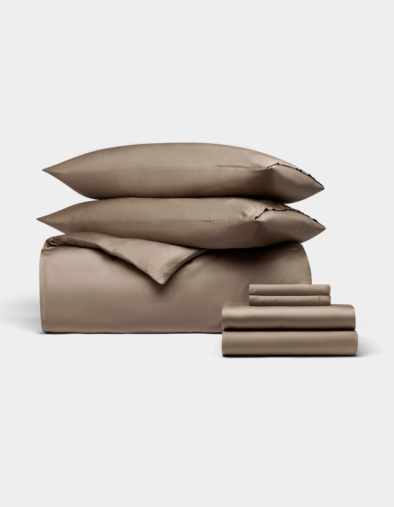 The Bamboo Bedding Ultimate Bundle by Cozy Earth features taupe-colored sheets, a fitted sheet, flat sheet, pillowcases, and two stacked pillows, all neatly arranged against a plain white background.