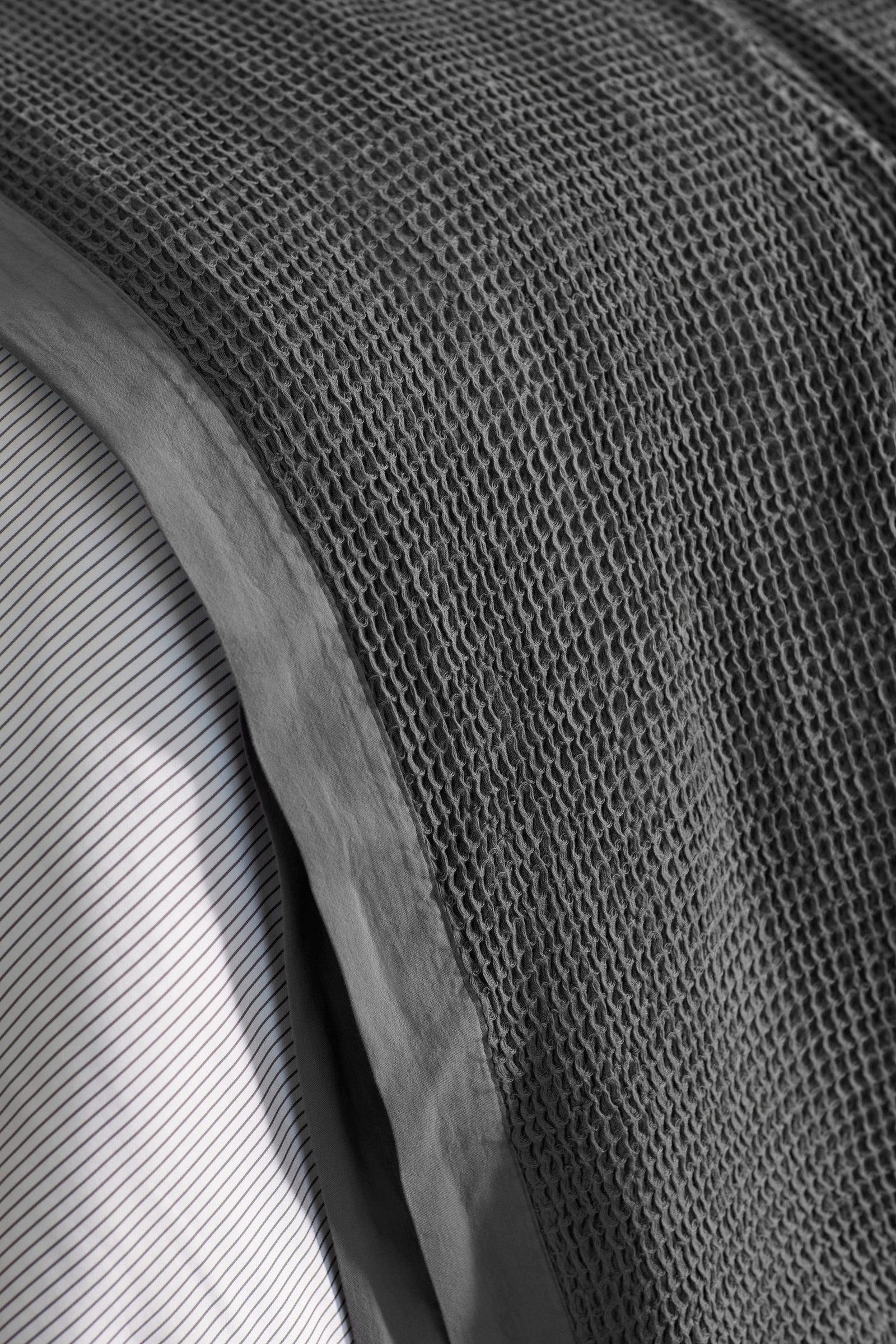 Close-up of the Cozy Earth Waffle Blanket, featuring a textured dark gray waffle weave, layered over a white sheet with thin black stripes.