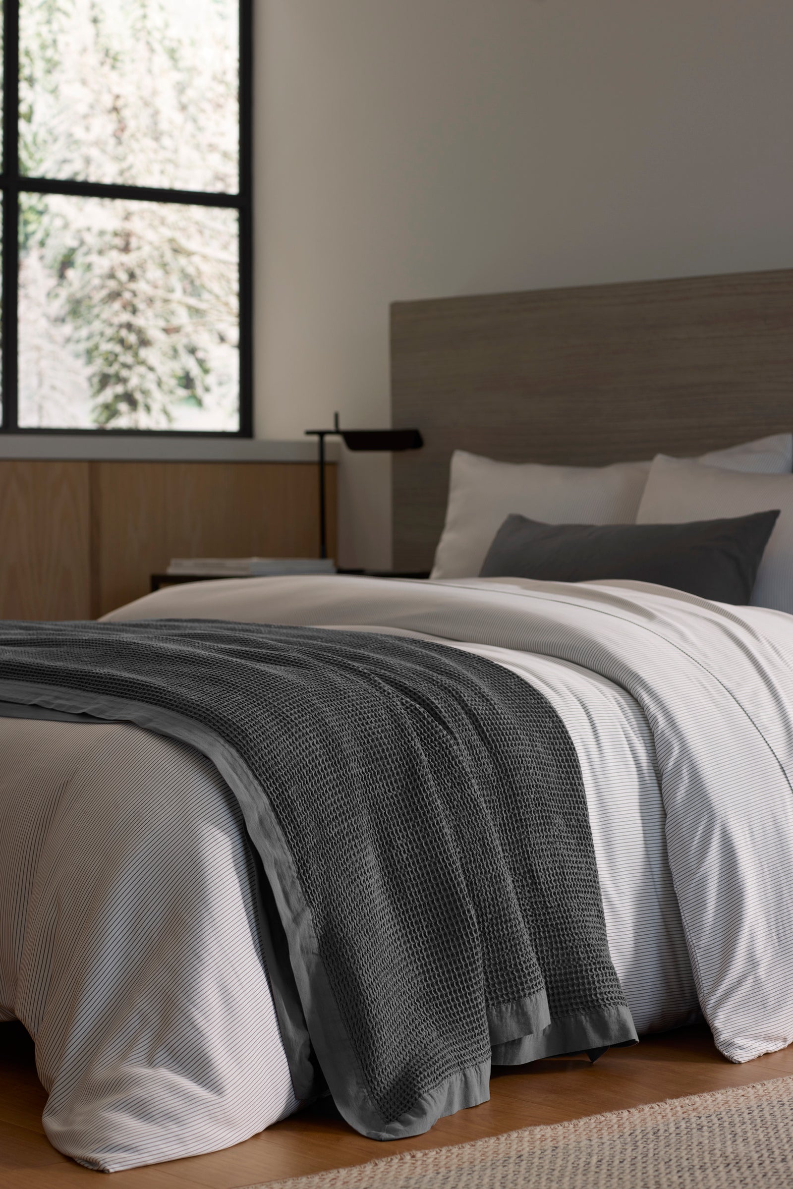 A modern bedroom features a neatly made bed with white linens and the Cozy Earth Waffle Blanket in dark gray, complemented by light wood accents and a large window filling the space with natural light.