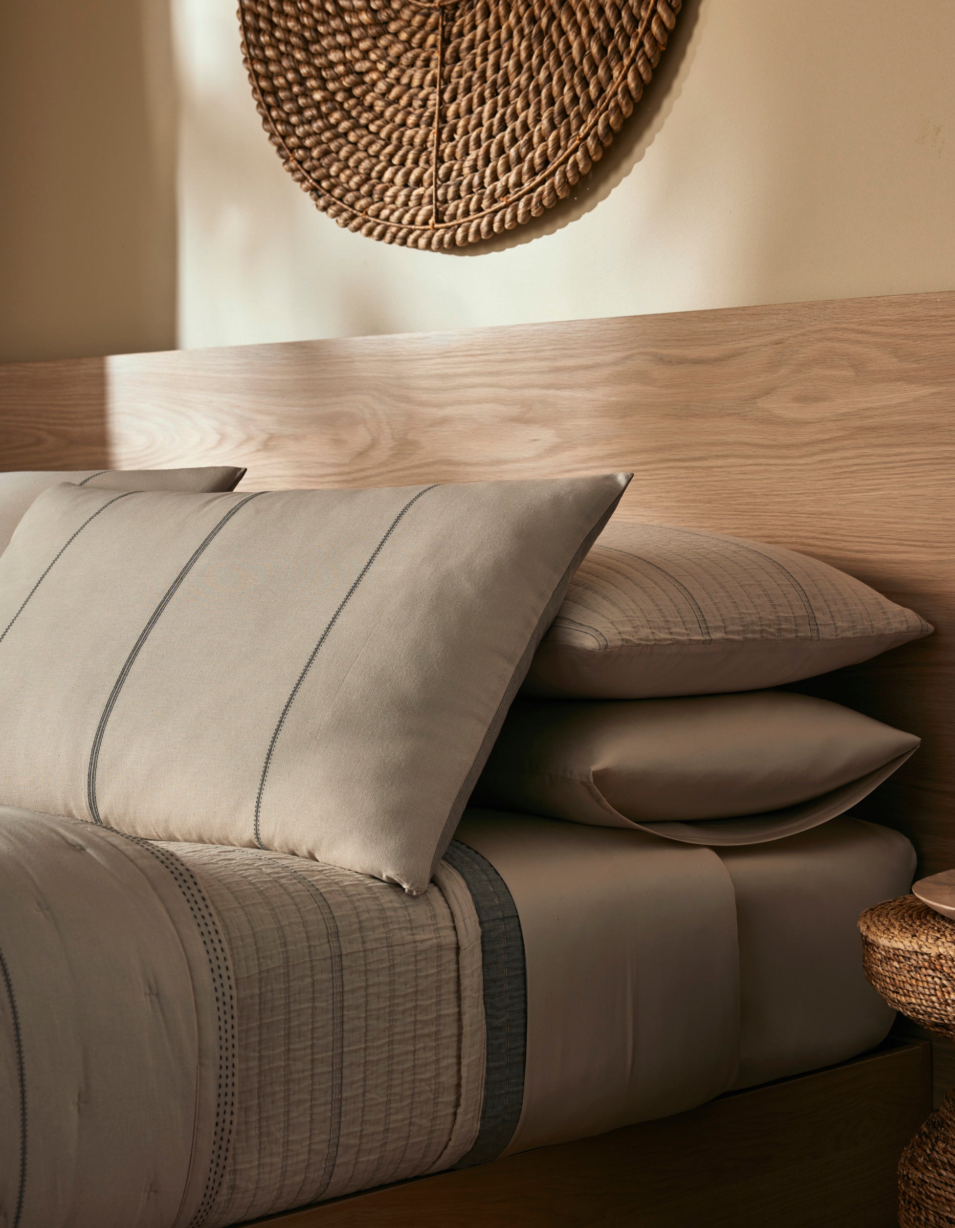 A cozy bedroom featuring the Cozy Earth Baja Double Sided Sham with taupe and gray bedding, stacked pillows, a light wood headboard, and a woven wall piece above, all softly lit for a warm ambiance.