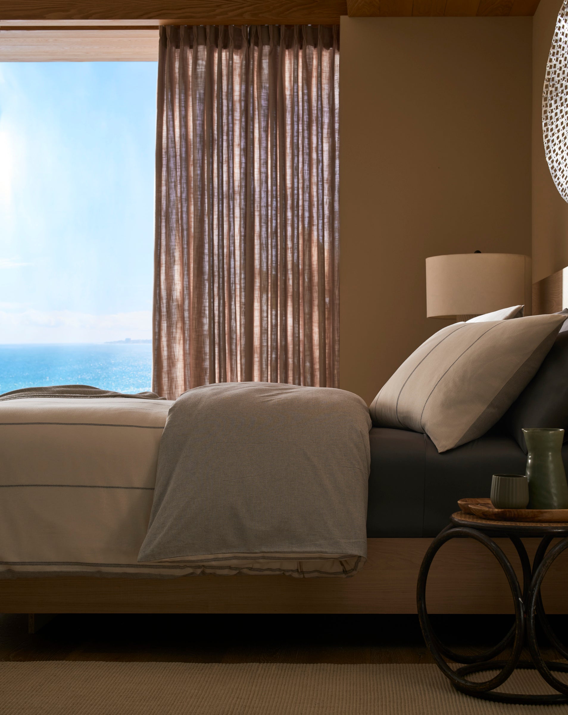 A sunlit bedroom features a neatly made bed with the Baja Duvet Cover by Cozy Earth, neutral bedding, and a large window with sheer curtains overlooking the ocean. A bedside table holds a green vase, cup, and book.|Color:Coal Stripe/Striped Alabaster