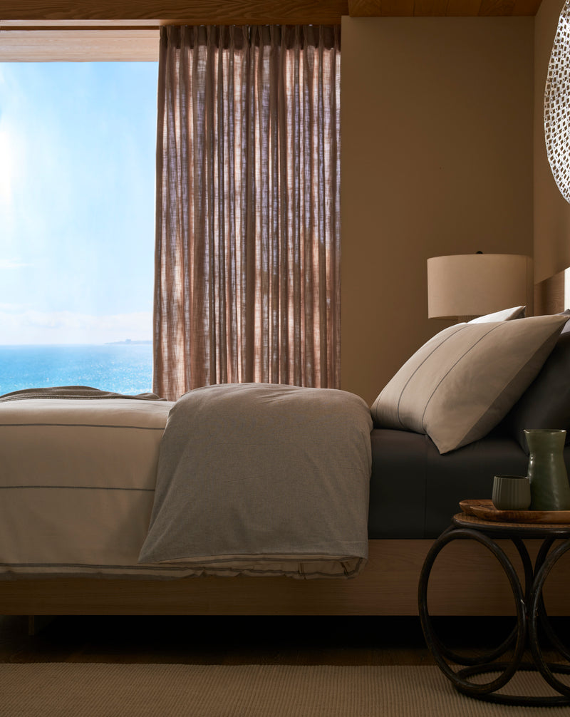 A sunlit bedroom features a neatly made bed with the Baja Duvet Cover by Cozy Earth, neutral bedding, and a large window with sheer curtains overlooking the ocean. A bedside table holds a green vase, cup, and book.