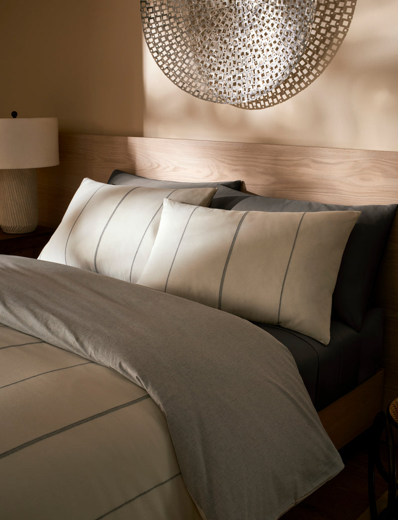 A bed made with the Cozy Earth Baja Duvet Cover in beige and gray stripes, two matching pillows, a wooden headboard, a bedside lamp, and a decorative metal wall hanging above the bed.