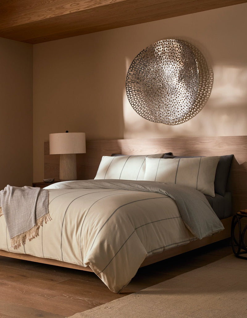 A modern bedroom features a low wooden bed with Cozy Earth's Baja Duvet Cover, beige and gray bedding, a textured throw, a bedside table with a lamp, and large metallic wall art above the bed. Warm neutral tones and soft natural light complete the space.
