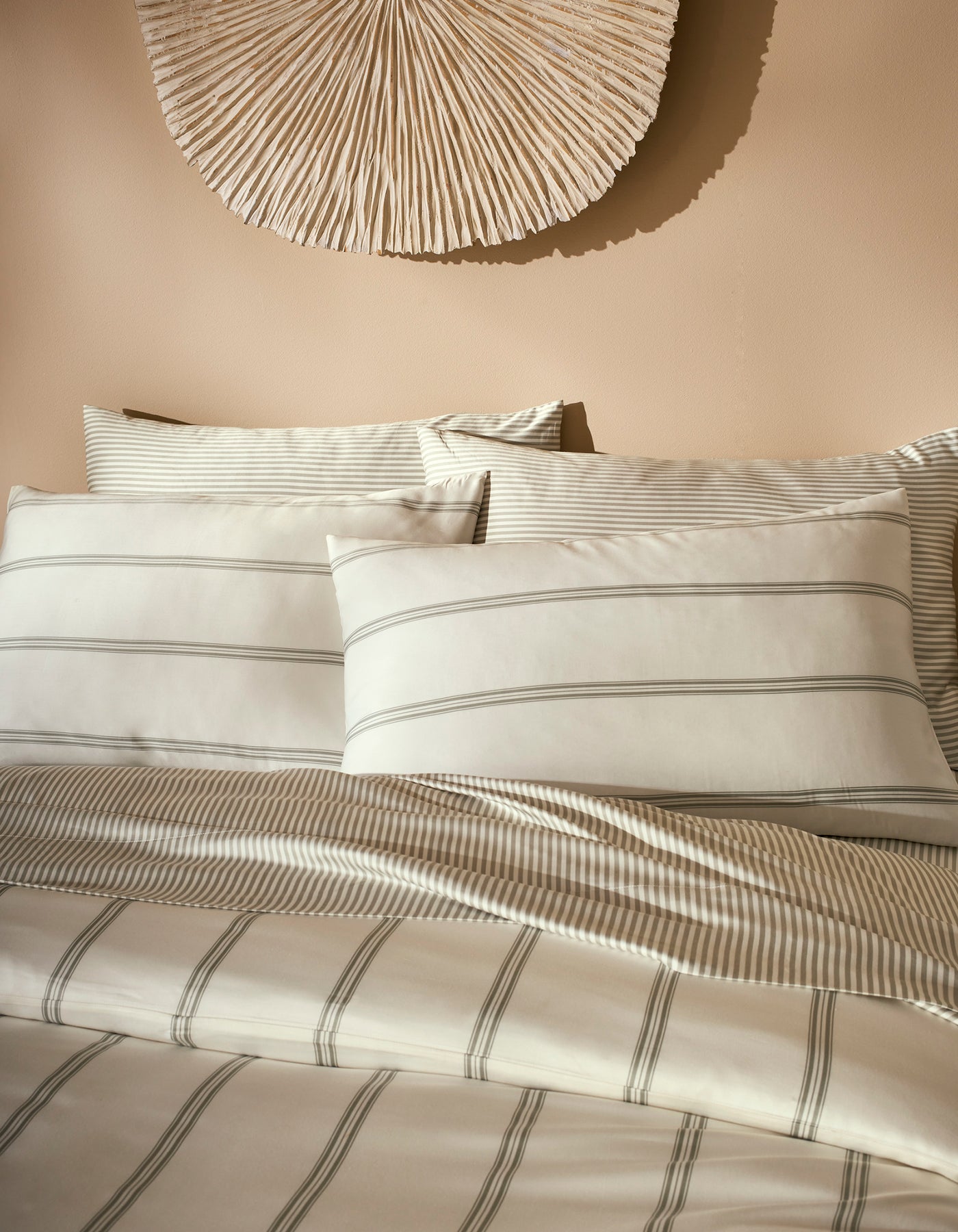 A neatly made bed features Cozy Earth Bamboo Pillowcases in cream and light gray stripes, accented by matching bedding and set against a beige wall with a textured, fan-shaped decoration above the headboard.