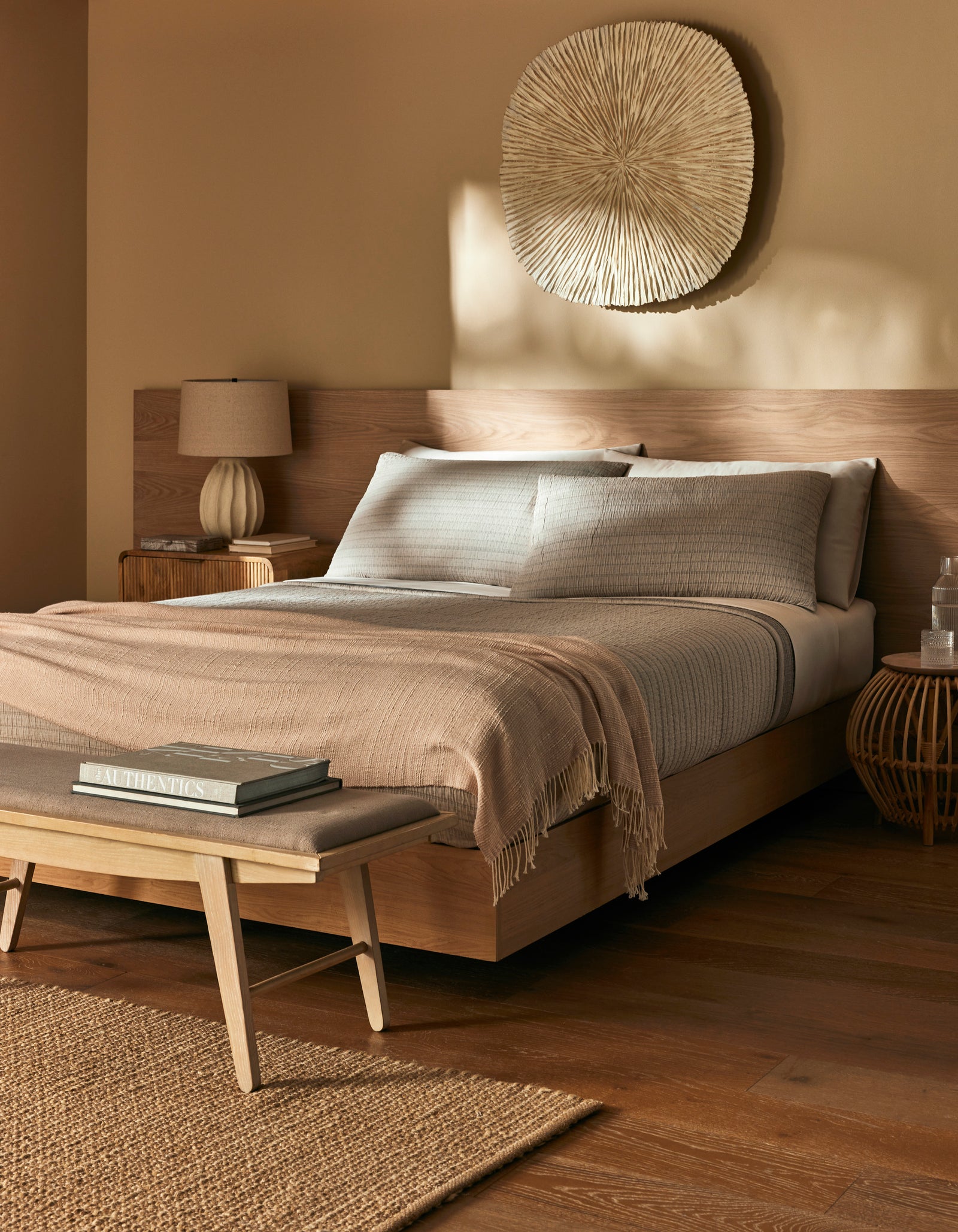 A cozy bedroom featuring a wooden bed frame, the Baja Matelasse Coverlet by Cozy Earth, a textured throw, bedside table with lamp, woven wall art, bench at the foot of the bed, and warm natural lighting.