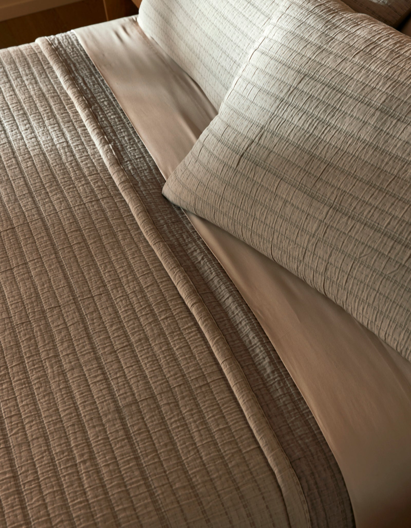 Close-up of a neatly made bed featuring the Cozy Earth Baja Matelasse Coverlet in light gray with matching pillows, accented by a smooth white sheet underneath. The bedding looks clean and inviting.