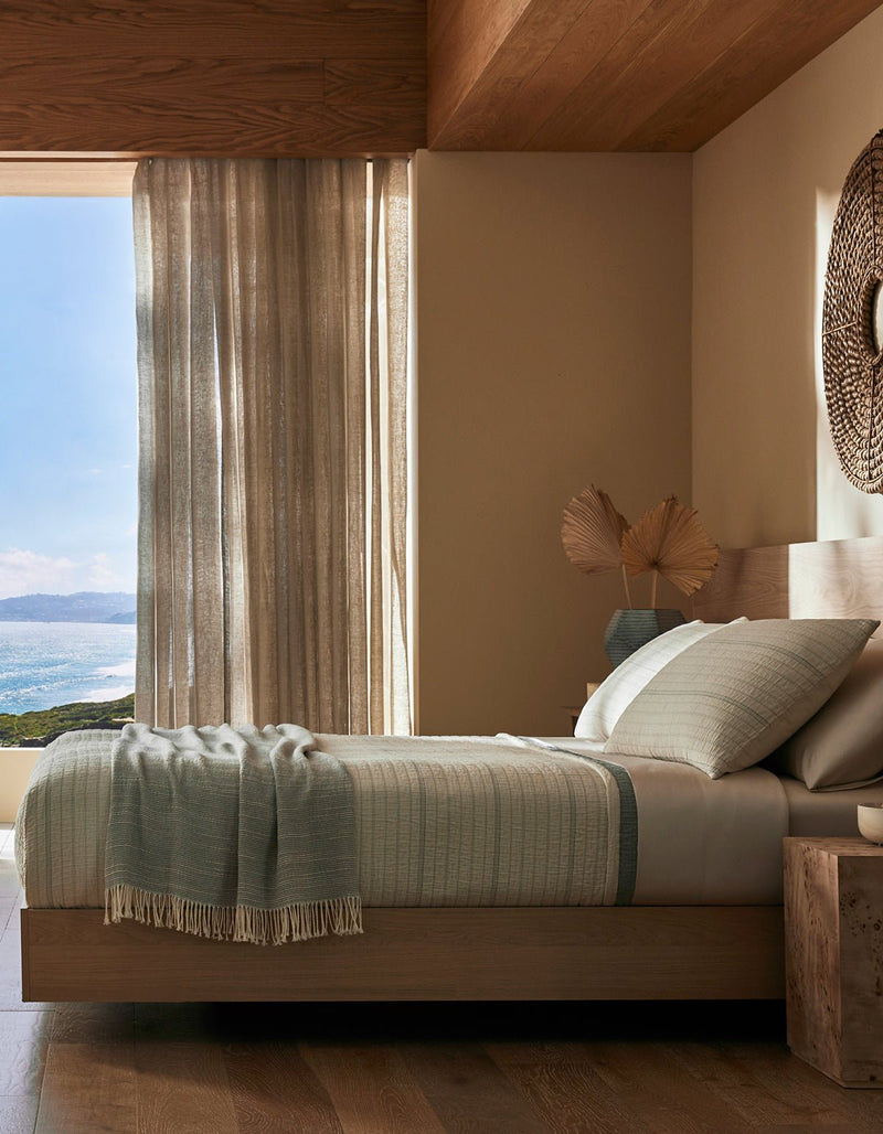 A serene bedroom features the Cozy Earth Baja Matelasse Bundle, soft neutral bedding, and decorative pillows. Light beige curtains are partially open to an ocean view, with natural light filling this cozy, minimalist space.
