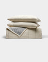 The Cozy Earth Baja Matelasse Bundle features two beige pillows stacked on a folded beige quilt with a gray inner lining, all set against a white background.|Color:Alabaster/Oceanside