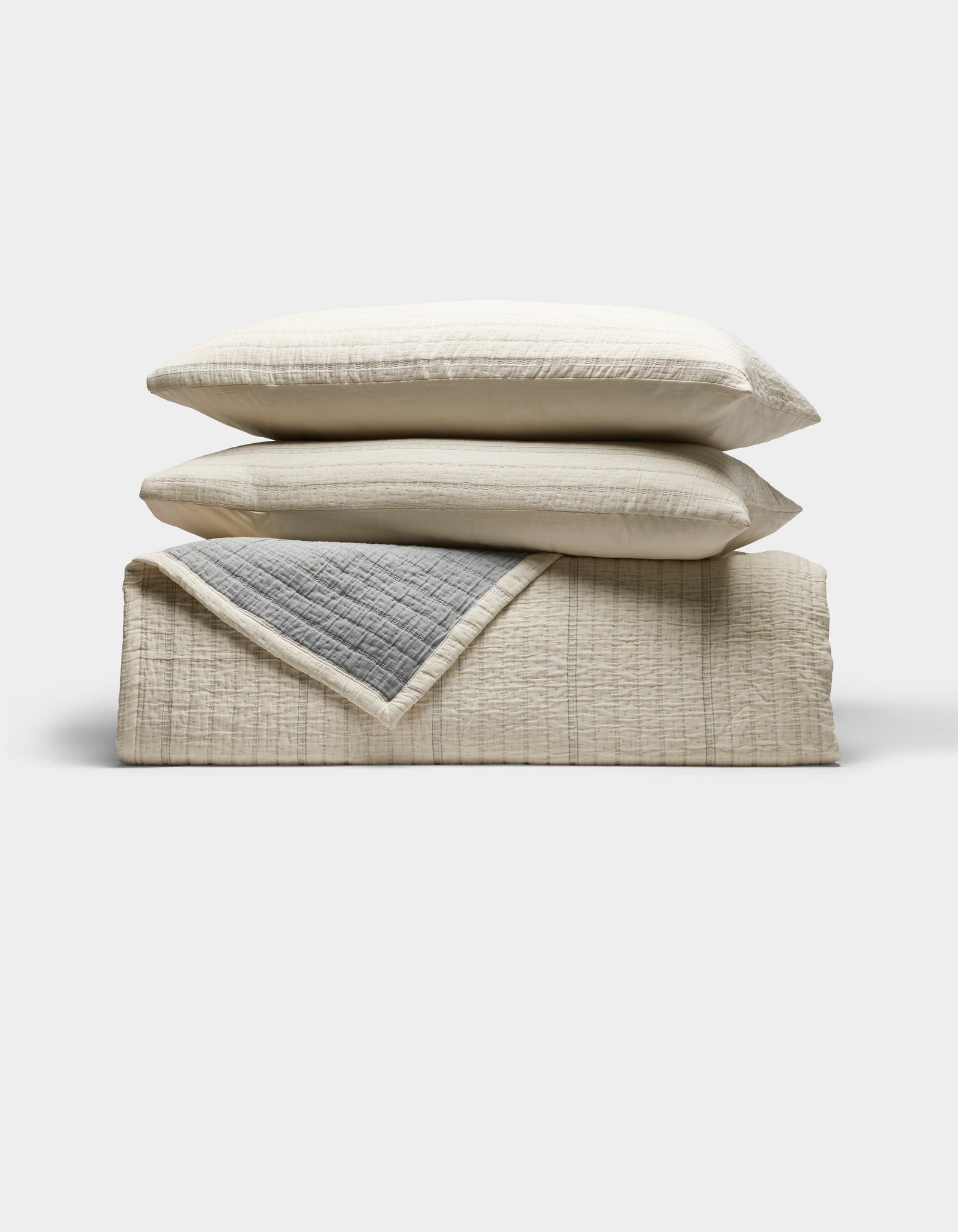 The Cozy Earth Baja Matelasse Bundle features two beige pillows stacked on a folded beige quilt with a gray inner lining, all set against a white background.