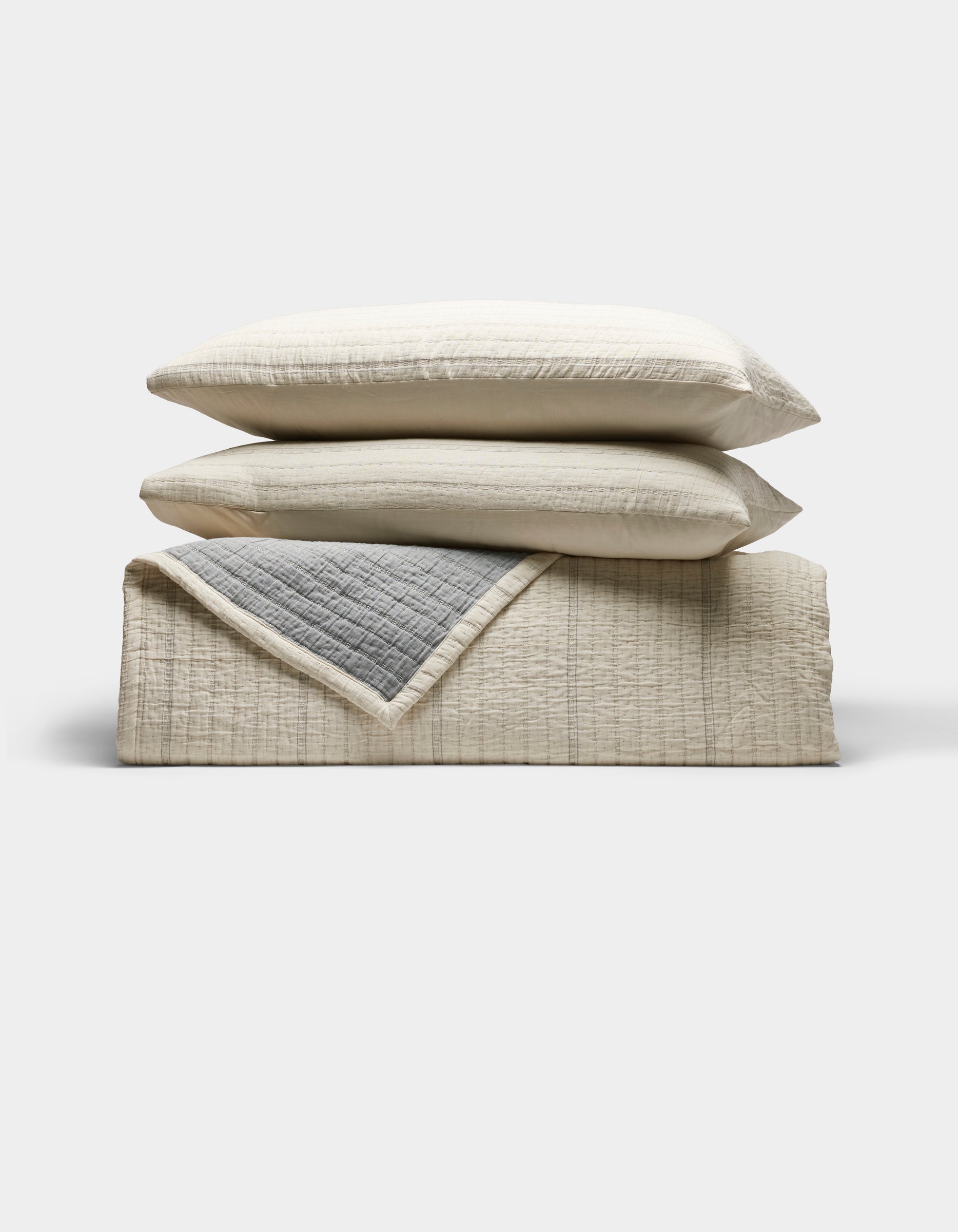 The Cozy Earth Baja Matelasse Bundle features two beige pillows stacked on a folded beige quilt with a gray inner lining, all set against a white background.|Color:Alabaster/Oceanside