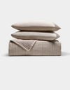 The Cozy Earth Baja Matelasse Bundle features two stacked, subtly striped pillows atop a folded, textured quilt in neutral beige tones, all set against a plain white background.|Color:Ebony/Taupe