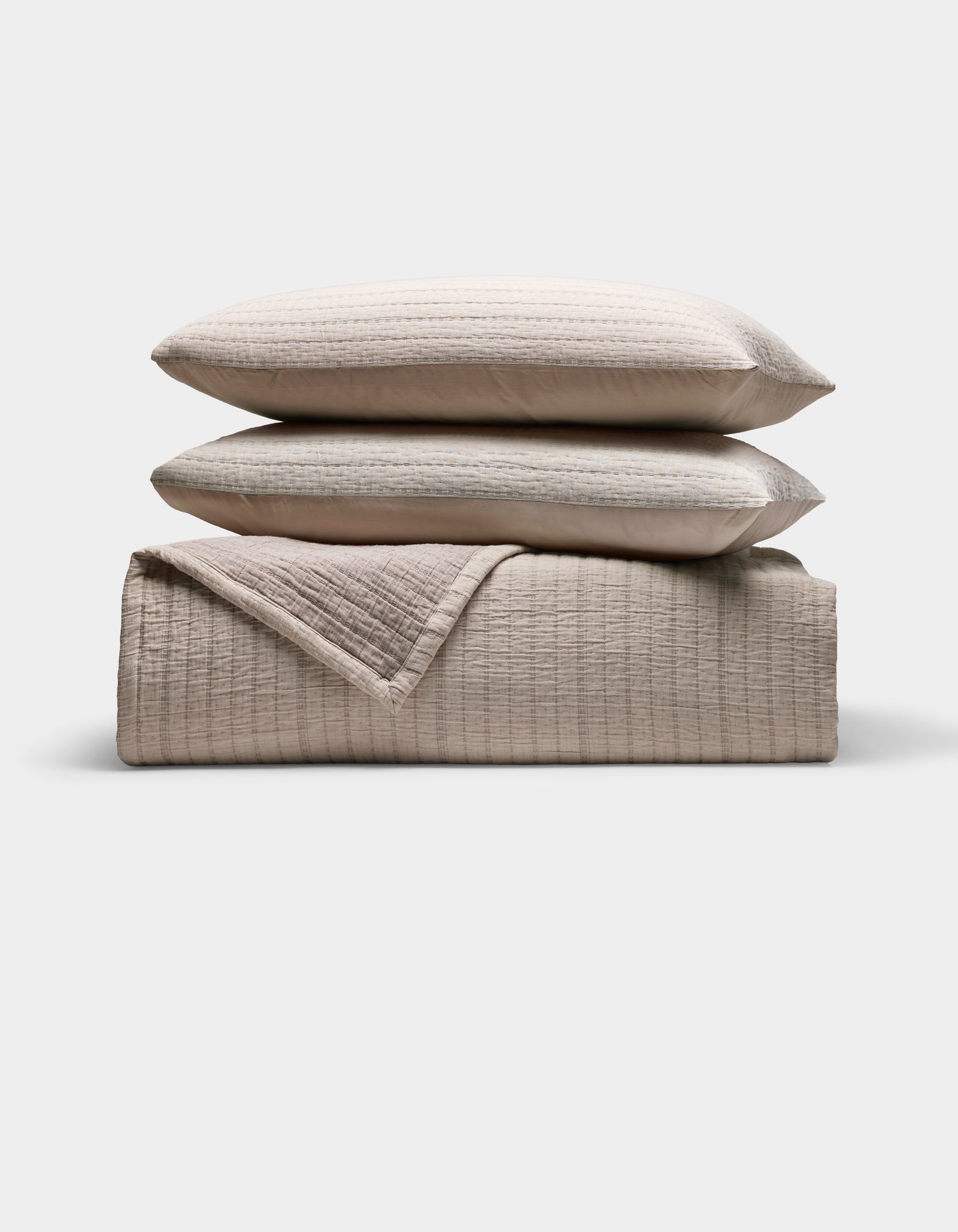 The Cozy Earth Baja Matelasse Bundle features two stacked, subtly striped pillows atop a folded, textured quilt in neutral beige tones, all set against a plain white background.