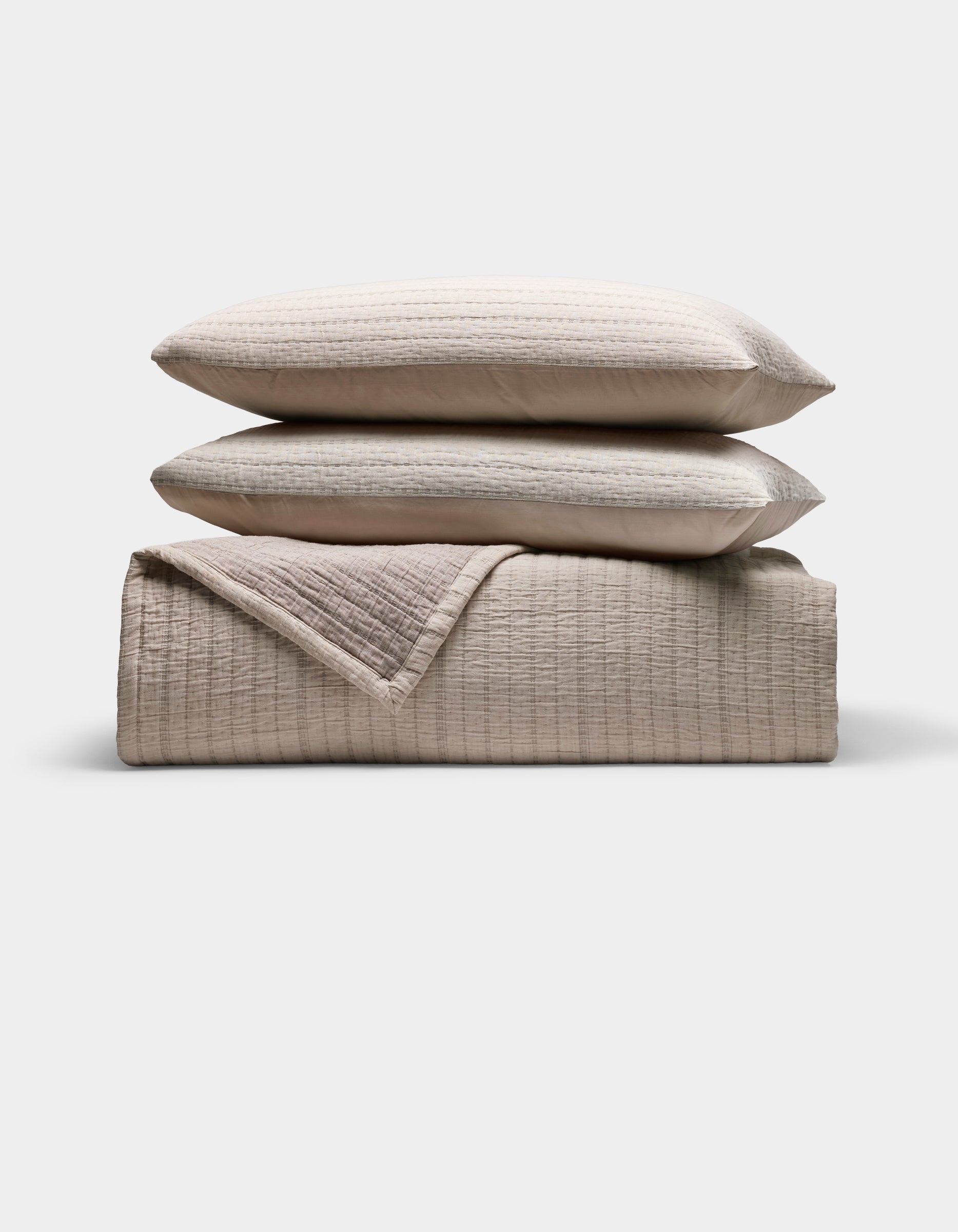 The Cozy Earth Baja Matelasse Bundle features two stacked, subtly striped pillows atop a folded, textured quilt in neutral beige tones, all set against a plain white background.|Color:Ebony/Taupe