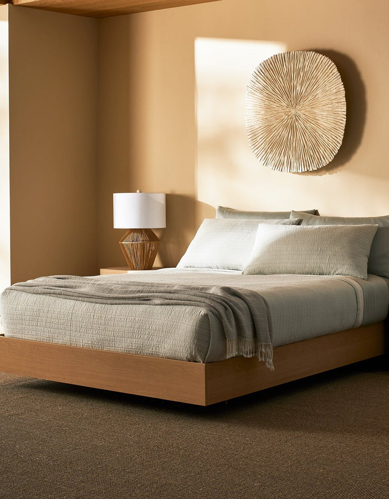 A modern bedroom featuring the Cozy Earth Baja Matelasse Bundle on a low wood bed, pale gray bedding with a gray throw, a wooden nightstand with lamp, and a decorative wall piece. The space showcases neutral, earthy tones.