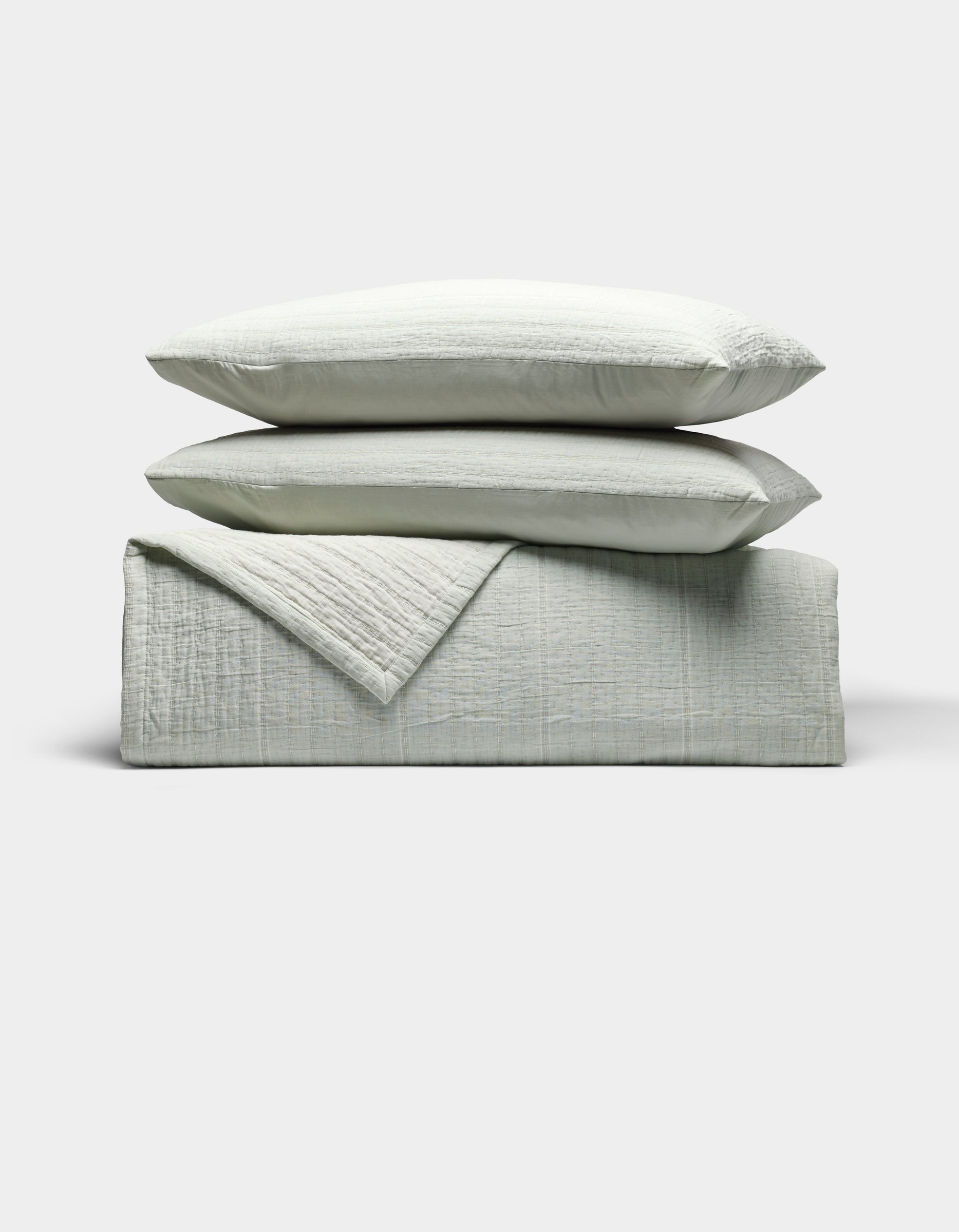 The Cozy Earth Baja Matelasse Bundle features two light gray pillows stacked atop a matching folded quilt, all set against a plain white background.|Color:Bone/Laurel