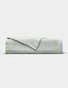 The Baja Matelasse Coverlet by Cozy Earth, in light gray with a textured finish, is neatly folded with one corner turned down and displayed against a plain white background.|Color:Bone/Laurel