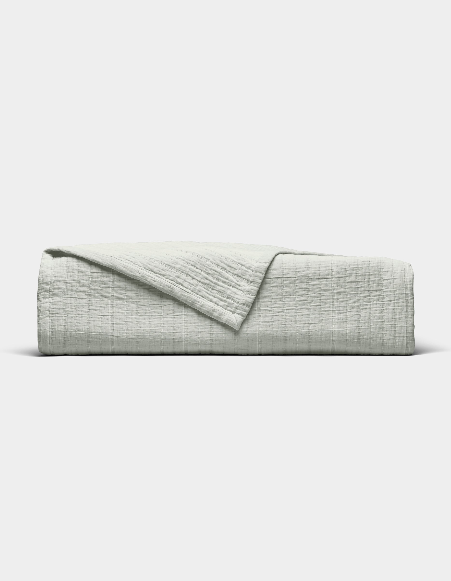 The Baja Matelasse Coverlet by Cozy Earth, in light gray with a textured finish, is neatly folded with one corner turned down and displayed against a plain white background.