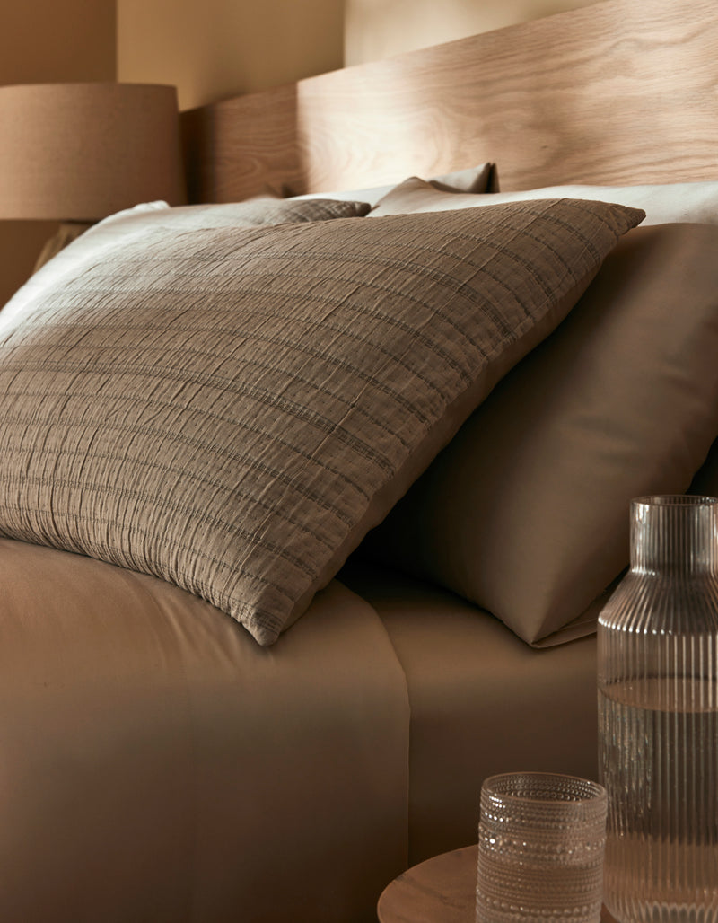 Close-up of a bed featuring Cozy Earth's Baja Matelasse Sham, with neutral bedding, pillows, a wooden headboard, and a glass carafe with tumbler on the bedside table. Warm lighting adds to the cozy atmosphere.