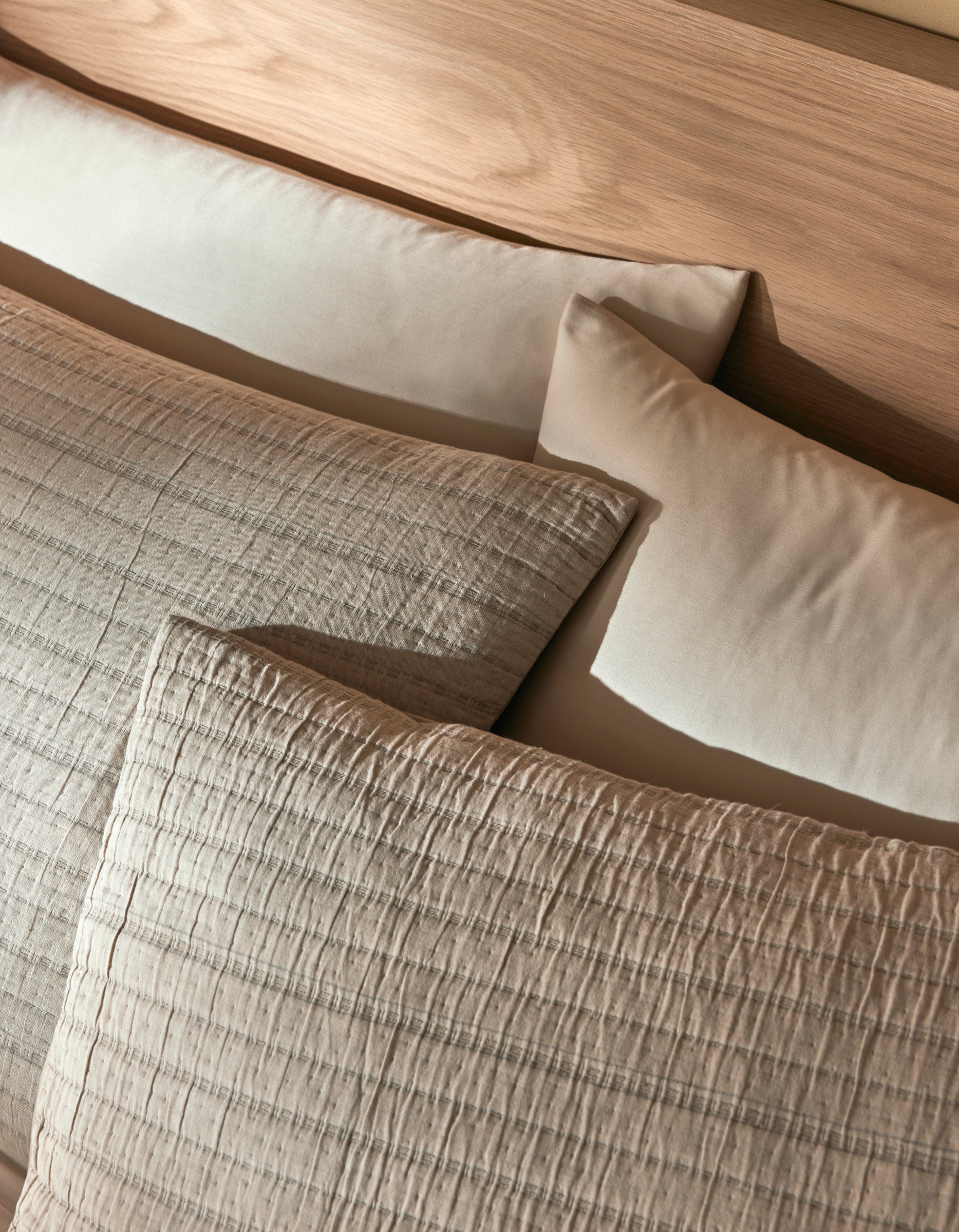 Two Cozy Earth Baja Matelasse Shams in beige and two plain cream pillows are arranged on a light wooden headboard, creating a cozy and inviting bed setting.