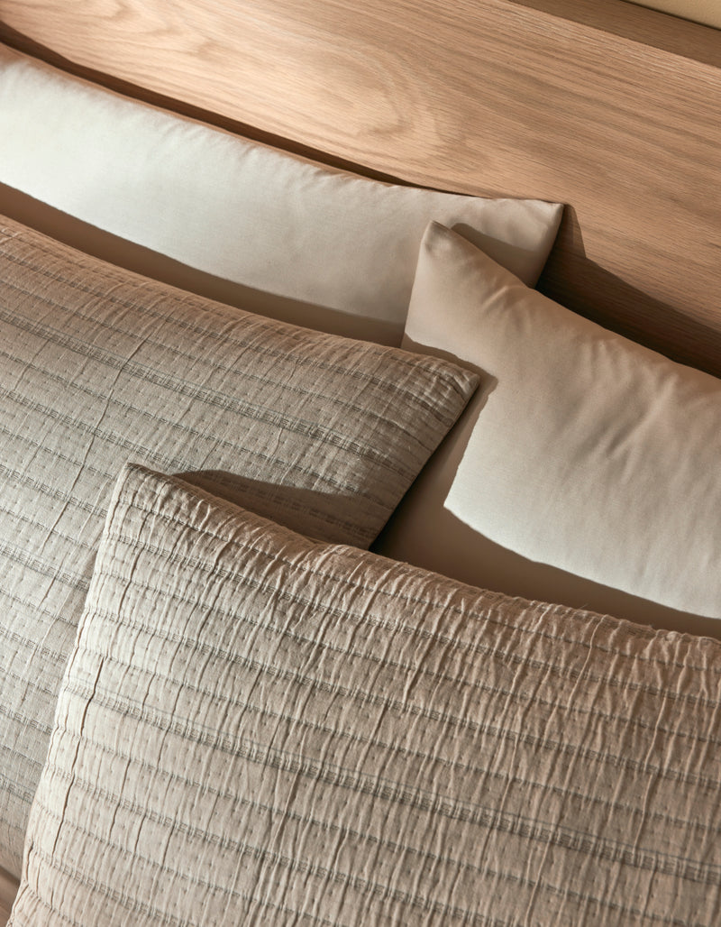 Two Cozy Earth Baja Matelasse Shams in beige and two plain cream pillows are arranged on a light wooden headboard, creating a cozy and inviting bed setting.