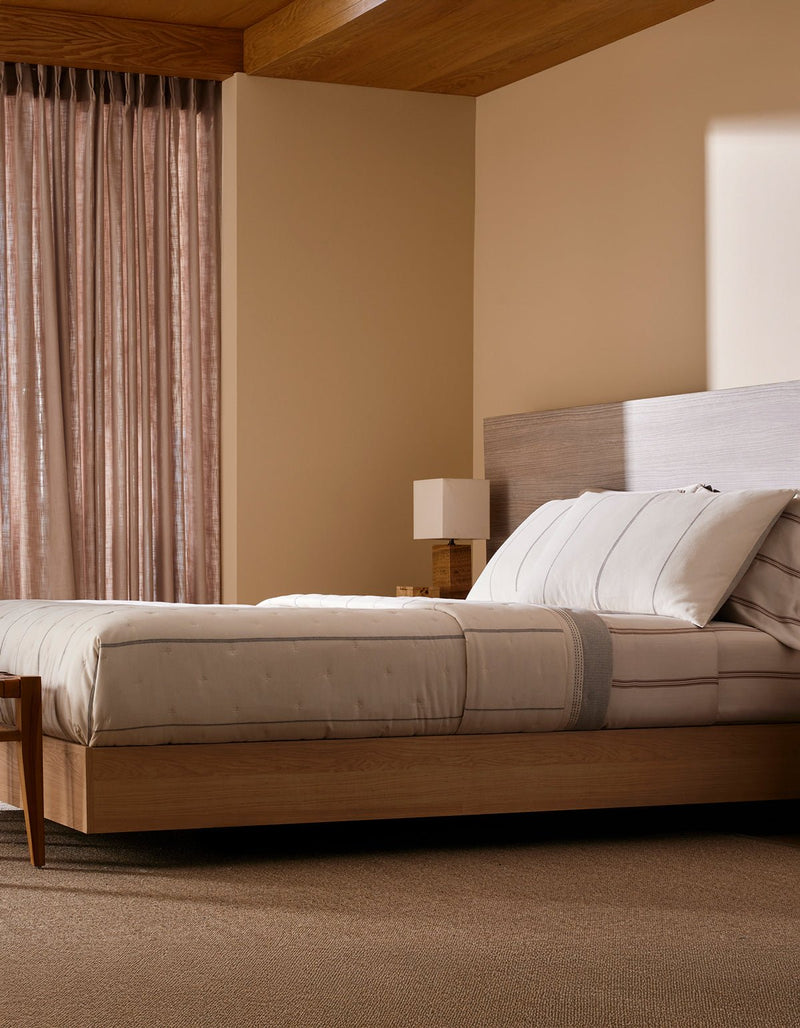 A modern bedroom features Cozy Earth’s Baja Quilt Bundle on a wooden platform bed, paired with a matching nightstand and lamp. Sheer curtains let in soft natural light, fostering a calm, minimalistic vibe.