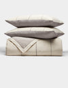 The Cozy Earth Baja Quilt Bundle features two cream pillows with gray edges stacked on a matching folded quilt, all with subtle stitched lines and a minimalist design, set against a plain white background.|Color:Coal Stripe/Striped Alabaster