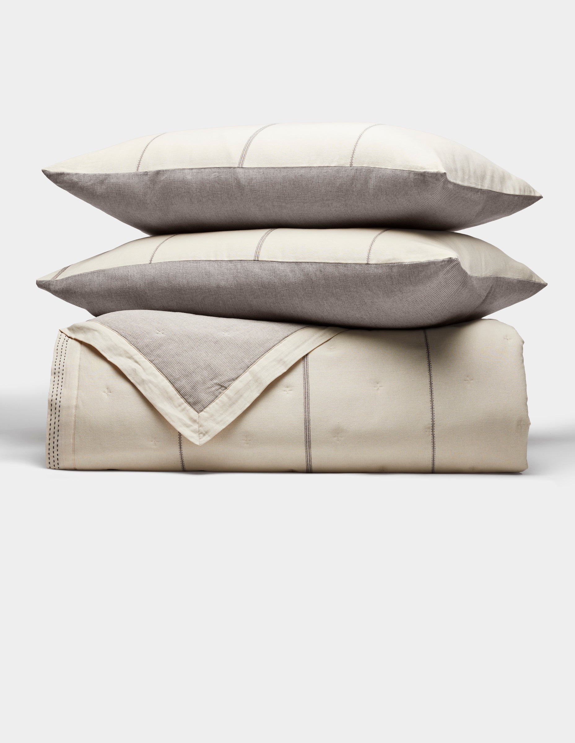 The Cozy Earth Baja Quilt Bundle features two cream pillows with gray edges stacked on a matching folded quilt, all with subtle stitched lines and a minimalist design, set against a plain white background.|Color:Coal Stripe/Striped Alabaster