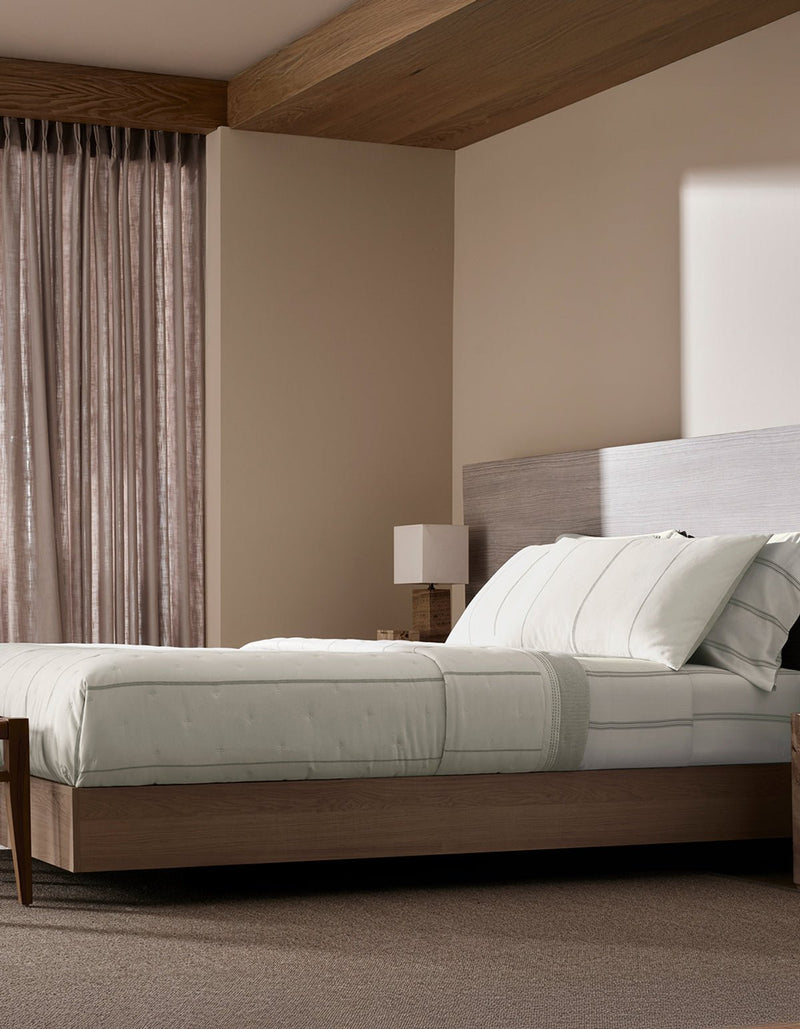 A modern bedroom with neutral tones features a wooden platform bed topped with the Cozy Earth Baja Quilt, light bedding, a minimalist nightstand with a square lamp, beige walls, and sheer curtains filtering sunlight that casts shadows on the wall.