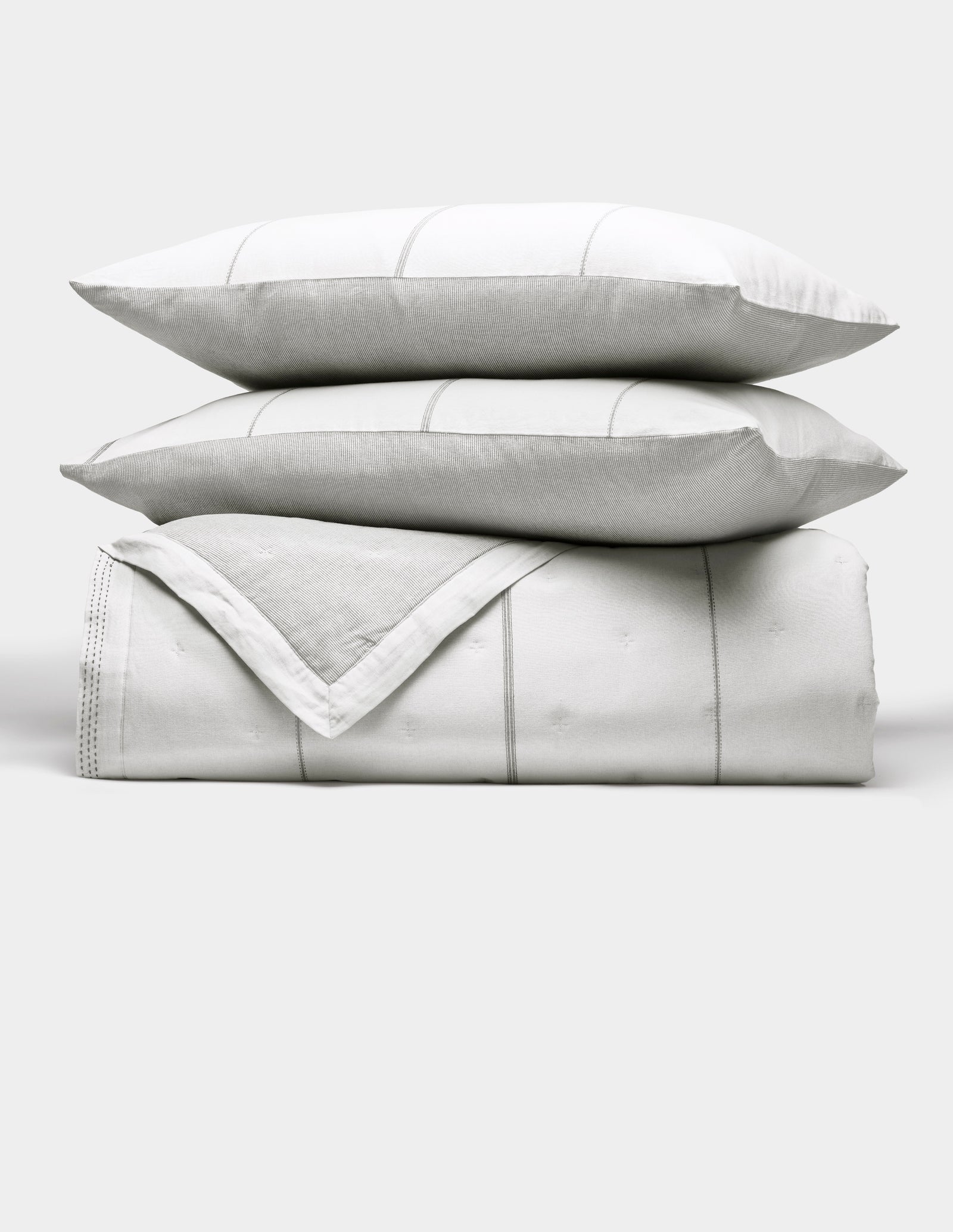 The Cozy Earth Baja Quilt Bundle features two white pillows with subtle gray stripes stacked atop a matching folded quilt, all set against a plain light background.