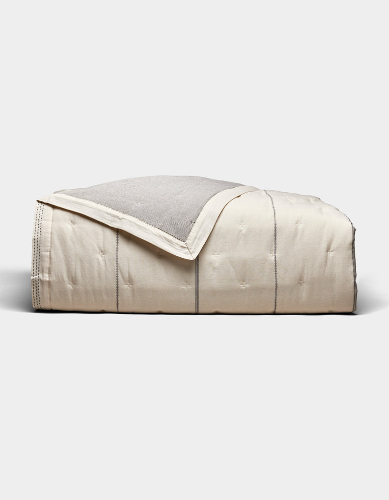 The Cozy Earth Baja Quilt, featuring a cream color with visible stitching and a light gray underside, is neatly folded and set against a plain white background.