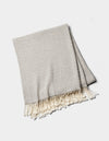 The Cozy Earth Baja Tassel Throw is a folded light gray woven blanket with subtle stripes and cream fringe on the ends, shown on a white background.|Color:Grey