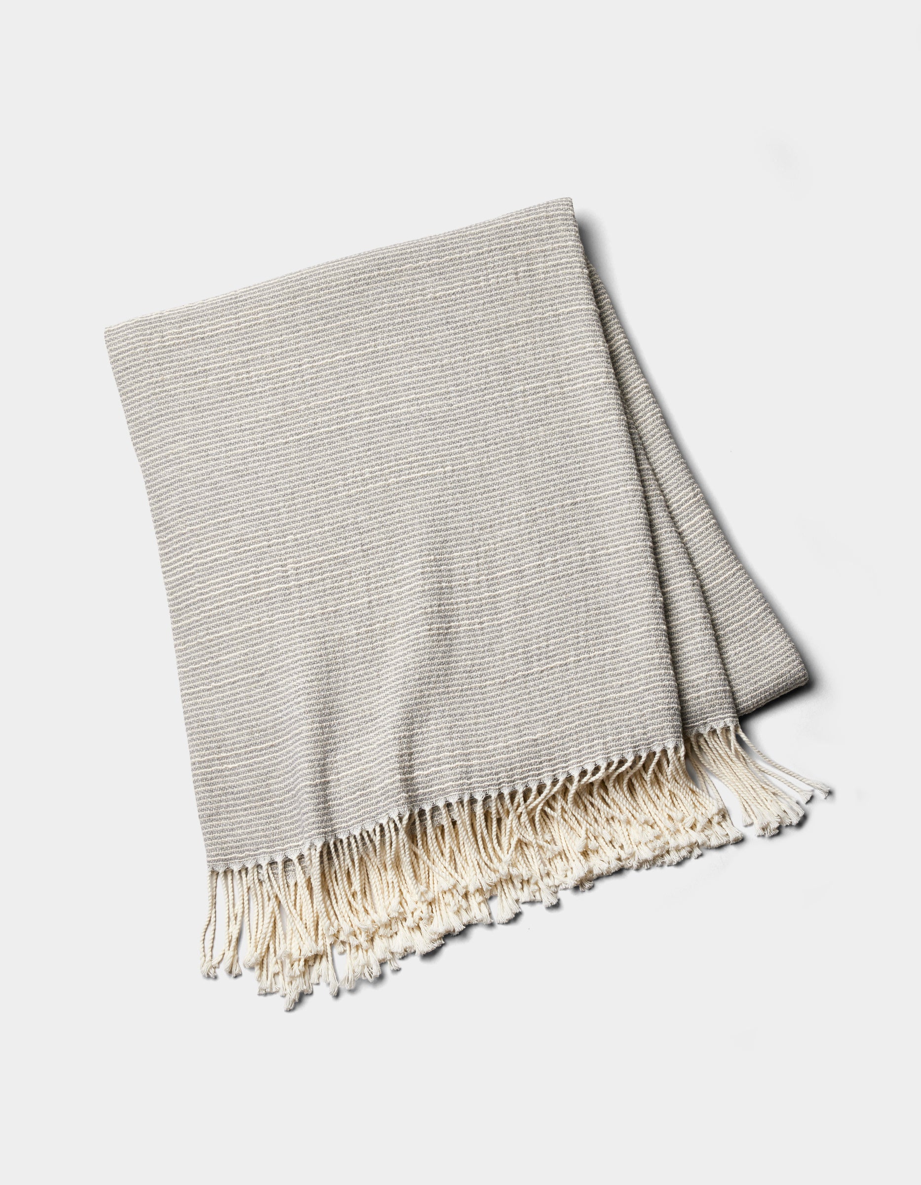 The Cozy Earth Baja Tassel Throw is a folded light gray woven blanket with subtle stripes and cream fringe on the ends, shown on a white background.|Color:Grey