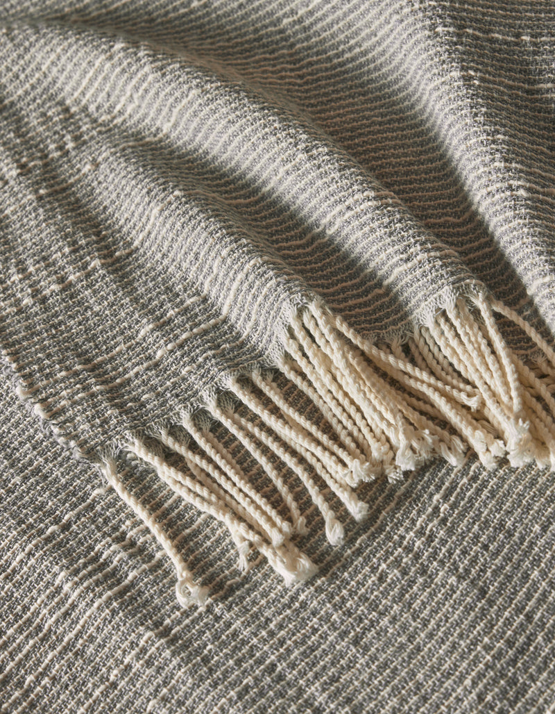The Baja Tassel Throw by Cozy Earth is shown in a close-up, highlighting its gray and white textured weave and fringed tassels draped elegantly to display its detailed pattern.