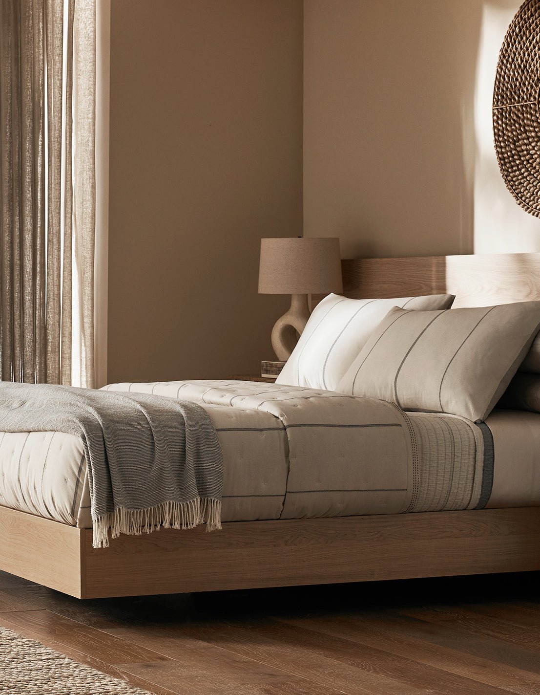 Minimalist bedroom featuring a light wood platform bed, beige bedding, the Cozy Earth Baja Tassel Throw in gray, round woven wall decor, a modern beige lamp, and soft natural light through sheer curtains.|Color:Grey
