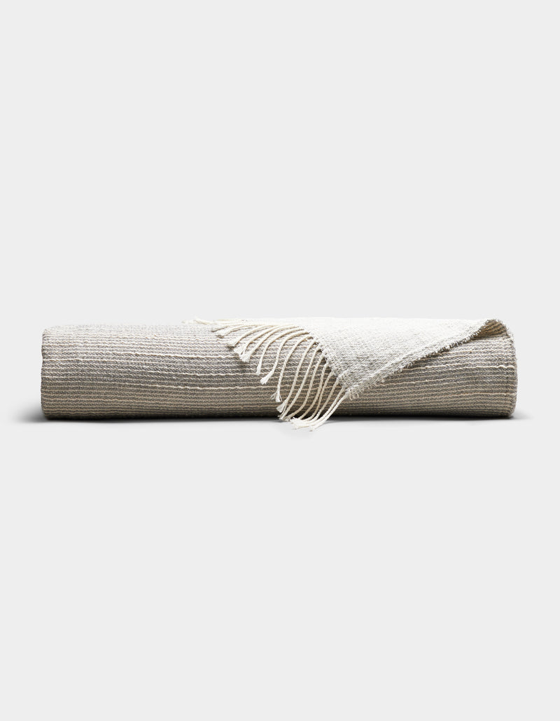 The Cozy Earth Baja Tassel Throw, a striped blanket in neutral beige and off-white tones with fringed edges, is neatly rolled and displayed on a plain, light background.