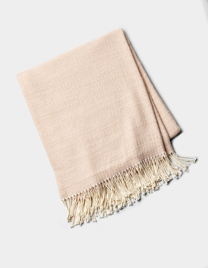 The Cozy Earth Baja Tassel Throw is a folded, light beige woven blanket with cream fringe on one end, shown on a plain white background.