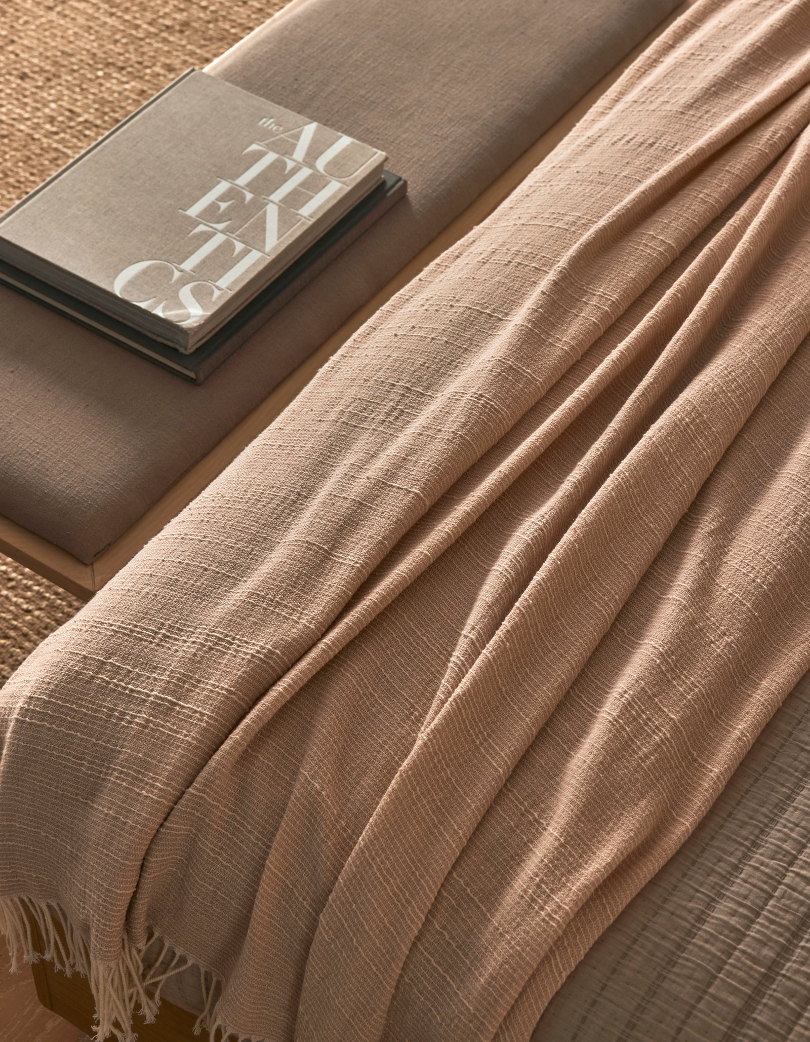The Baja Tassel Throw by Cozy Earth, in beige with a textured finish, is draped over a neutral-toned bench beside two stacked books (one titled "AUTHENTICS"), creating a cozy, minimalistic look.
