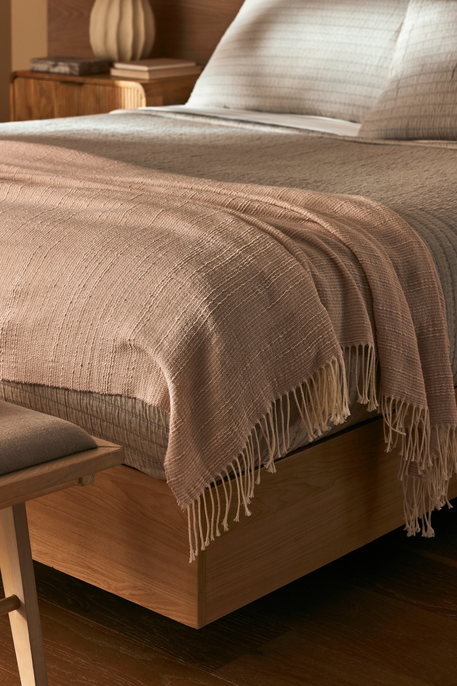 A close-up of a neatly made bed with the Cozy Earth Baja Tassel Throw draped at the foot. The wooden frame and neutral bedding create a cozy, minimalist vibe, complemented by the throw’s light texture.
