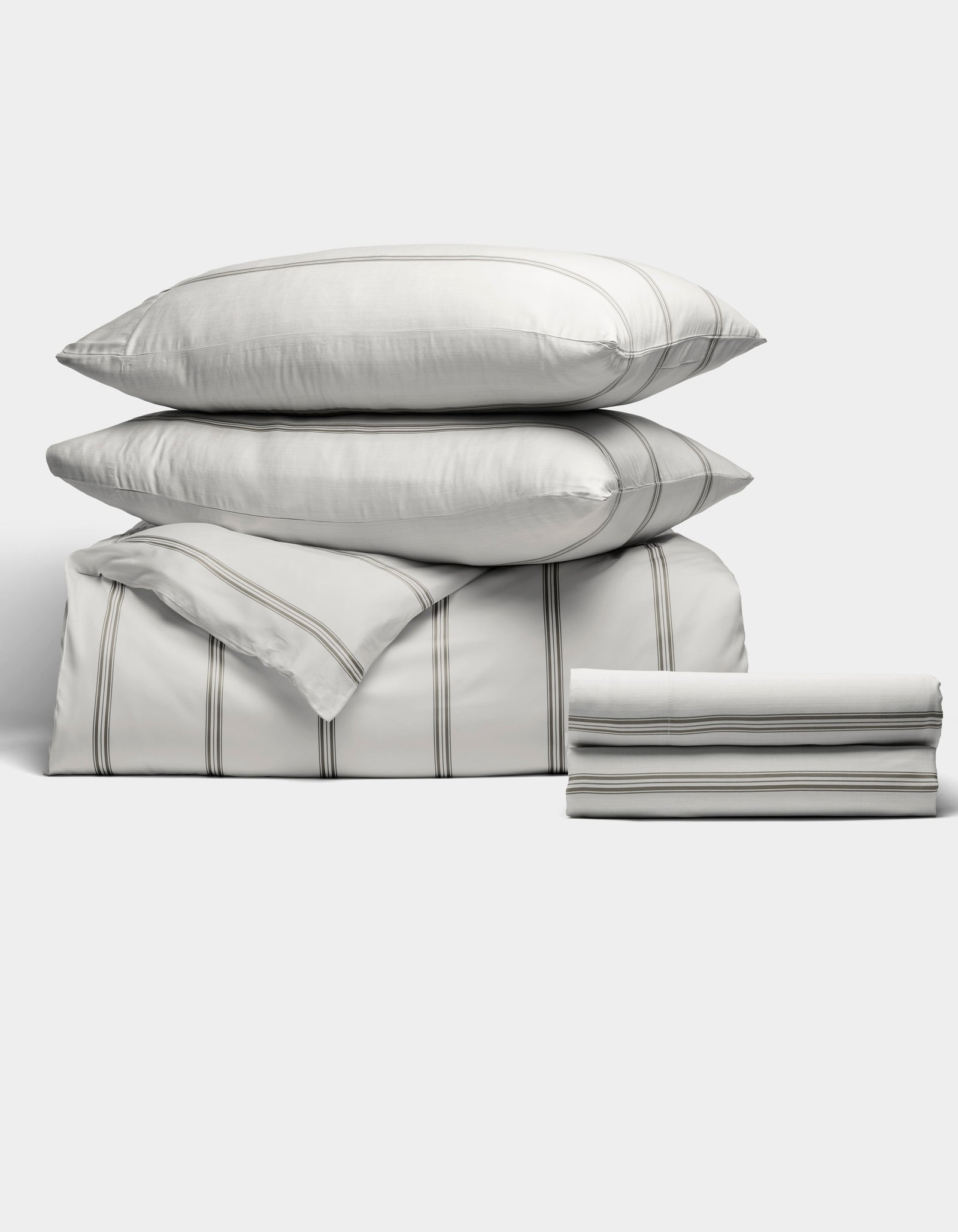 The Bamboo Bedding Deluxe Bundle by Cozy Earth features two pillows, a folded comforter, and a folded sheet in white with thin gray stripes, all neatly stacked against a plain light background.
