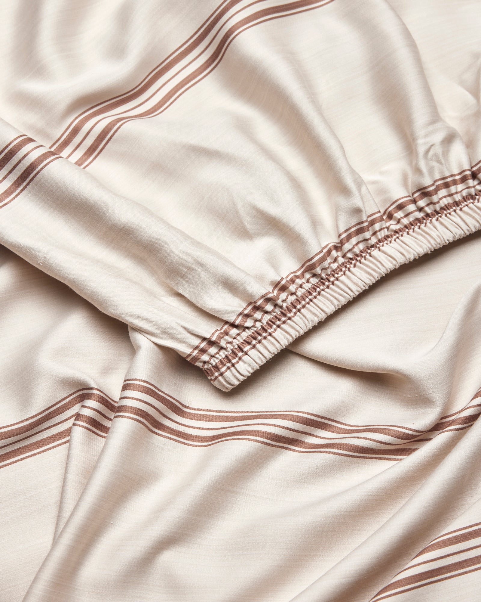 A close-up of Cozy Earth's Bamboo Fitted Sheet in beige, featuring brown horizontal stripes and an elastic gathered edge, displayed in soft folds.