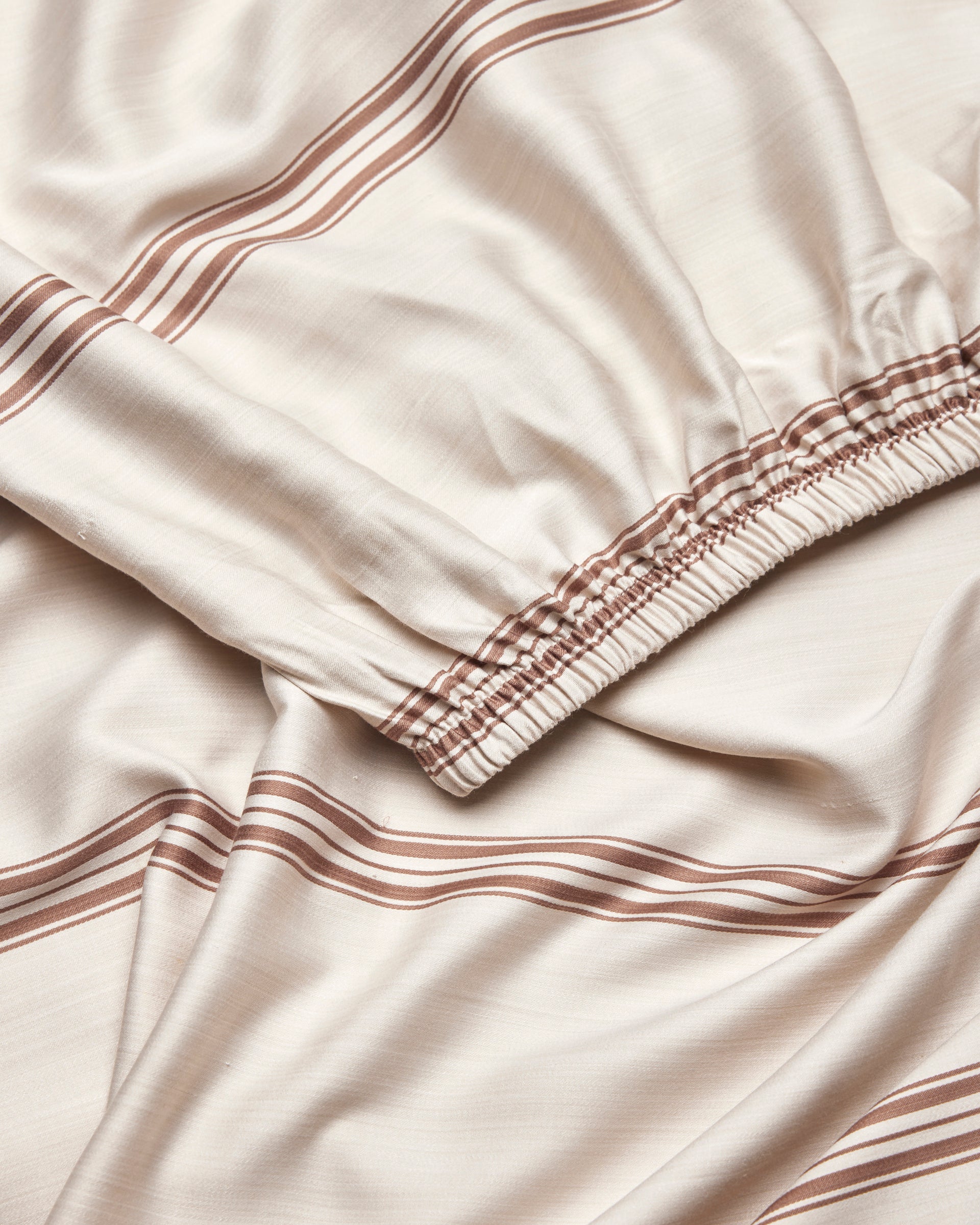 A close-up of Cozy Earth's Bamboo Fitted Sheet in beige, featuring brown horizontal stripes and an elastic gathered edge, displayed in soft folds.