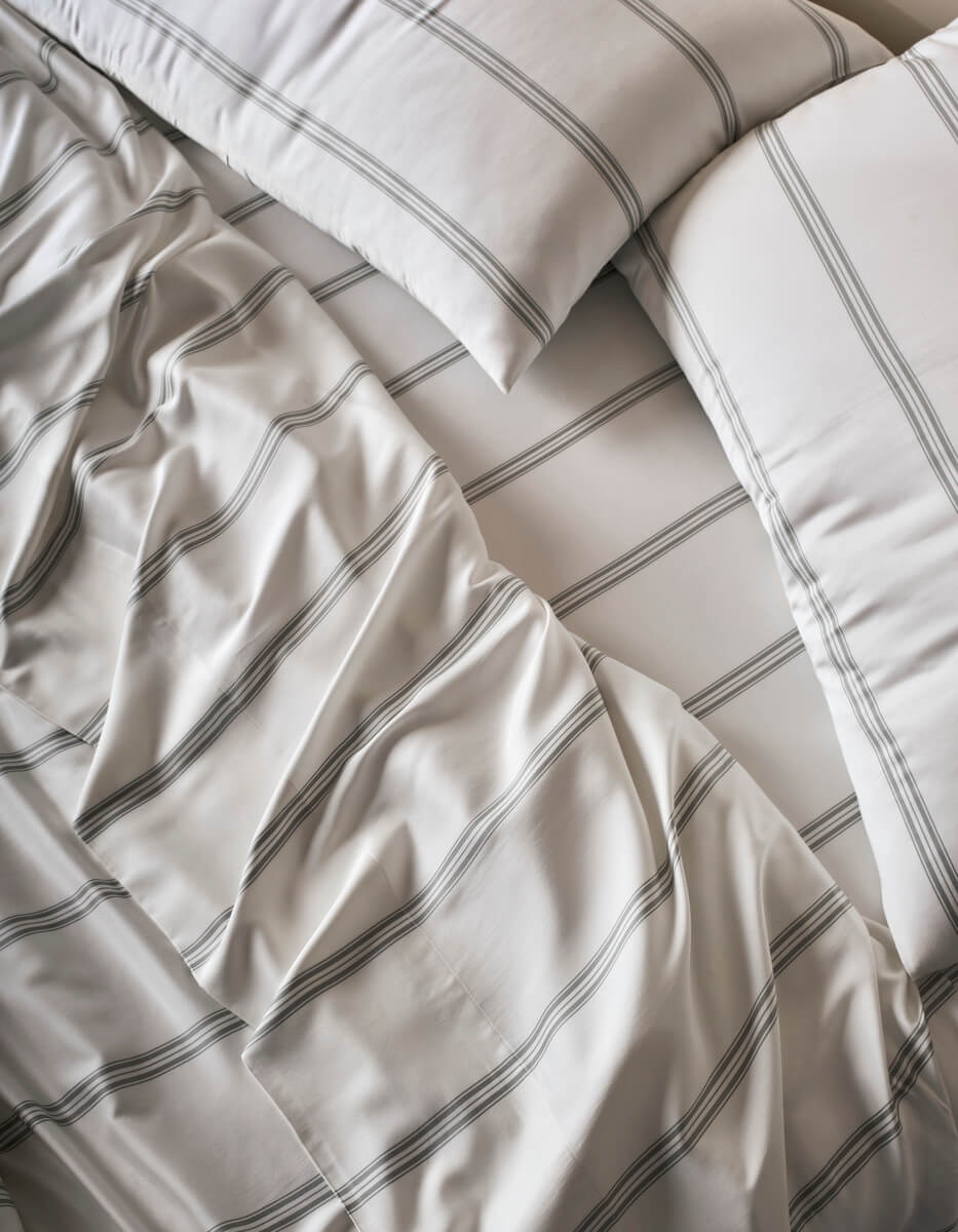 A close-up of a bed dressed in Cozy Earth’s Bamboo Sheet Set - Last Chance, with white sheets and pillowcases adorned with thin, gray stripes. The bedding is lightly rumpled, displaying gentle folds and creases in the fabric.