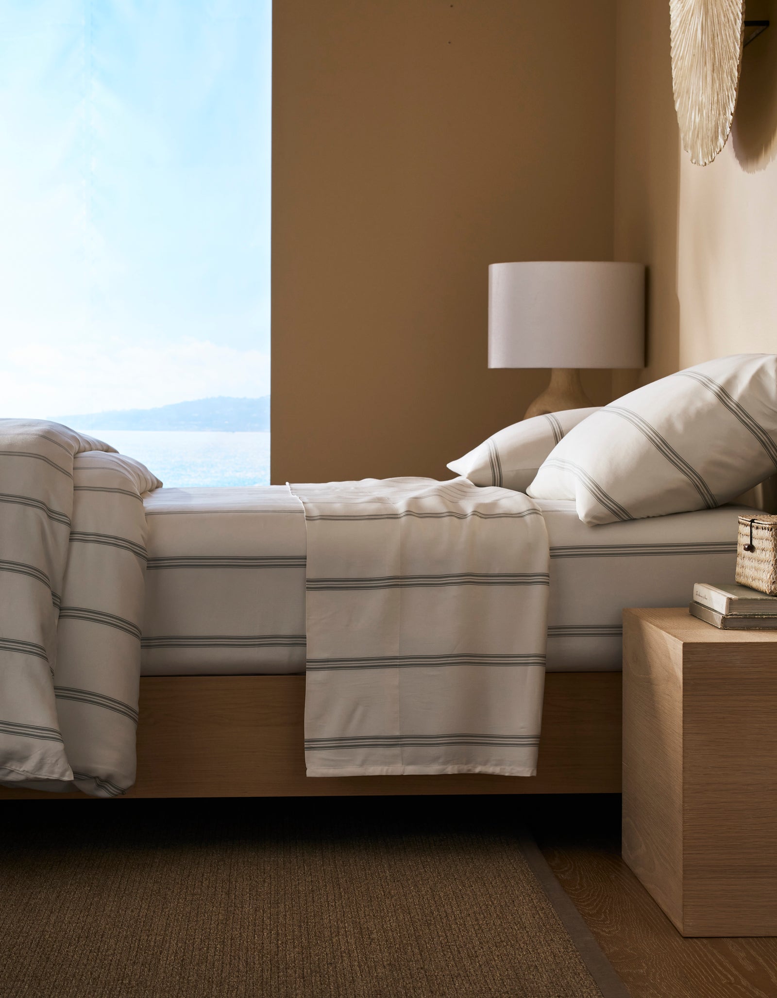 A modern bedroom featuring a neatly made bed with Cozy Earth’s Bamboo Sheet Set, a wooden nightstand with books and decor, a white lamp, and a large window offering views of water and sky.