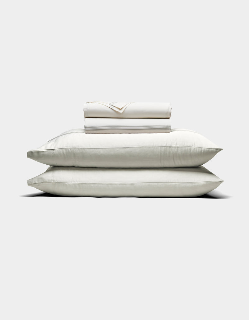 Two large white pillows are stacked with a folded Cozy Earth Bamboo Sheet Set and pillowcases placed on top, all arranged against a plain light background.