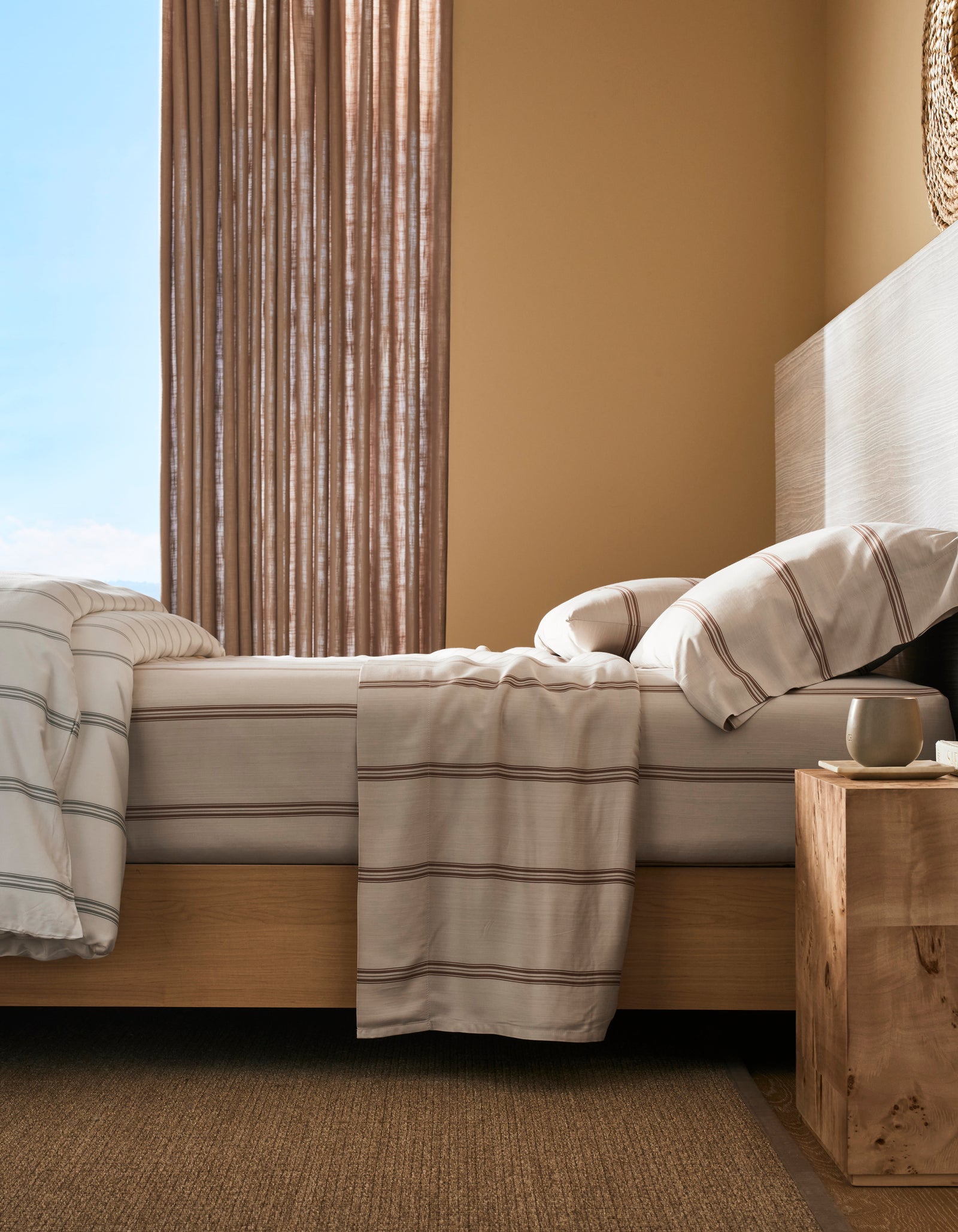 A neatly made bed features Cozy Earth's Bamboo Sheet Set with striped white and beige bedding, beside a wooden nightstand with a cup, in a sunlit room with beige walls and floor-length curtains.