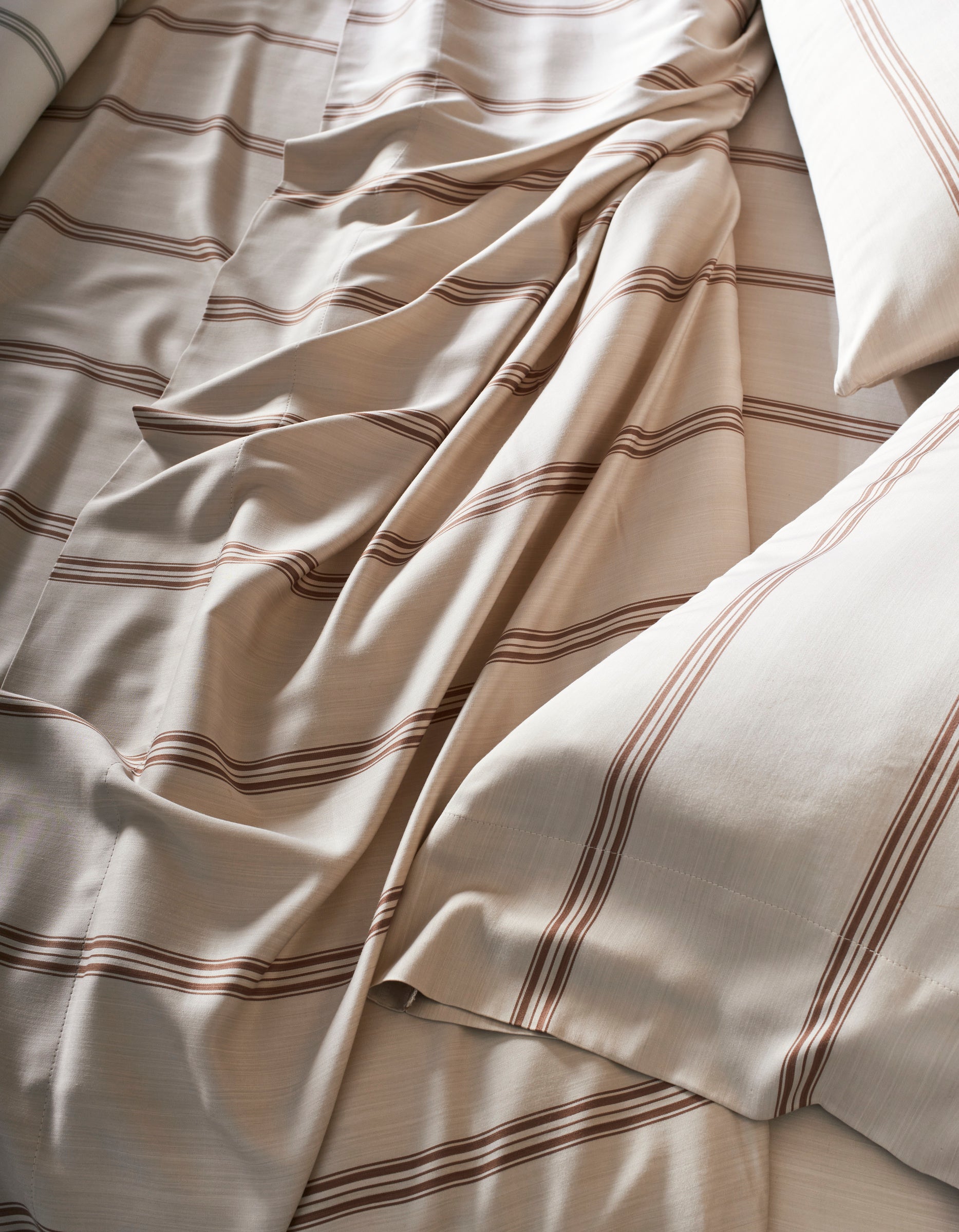 A close-up of the Cozy Earth Bamboo Sheet Set shows cream-colored sheets with brown horizontal stripes, slightly rumpled, and two pillows placed on either side.