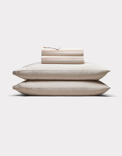 Two cream-colored pillows are stacked with Cozy Earth’s Bamboo Sheet Set, neatly folded on top, all arranged against a plain white background.|Color:Baja Stripe in Maple