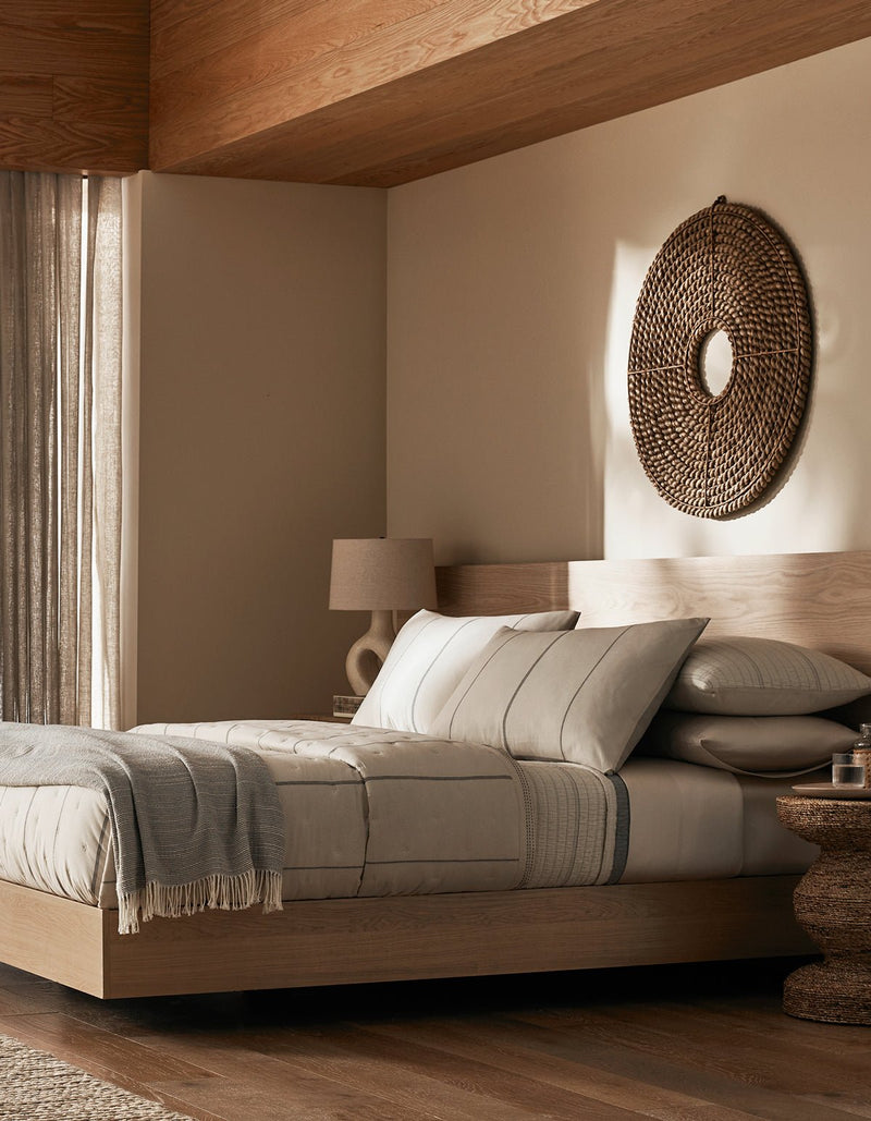 A modern, minimalist bedroom features a wooden bed frame with beige bedding, the Cozy Earth Baja Quilt, neutral pillows, a textured blanket, plus a woven circular wall decor, lamp, and round side table for a cozy sunlit look.